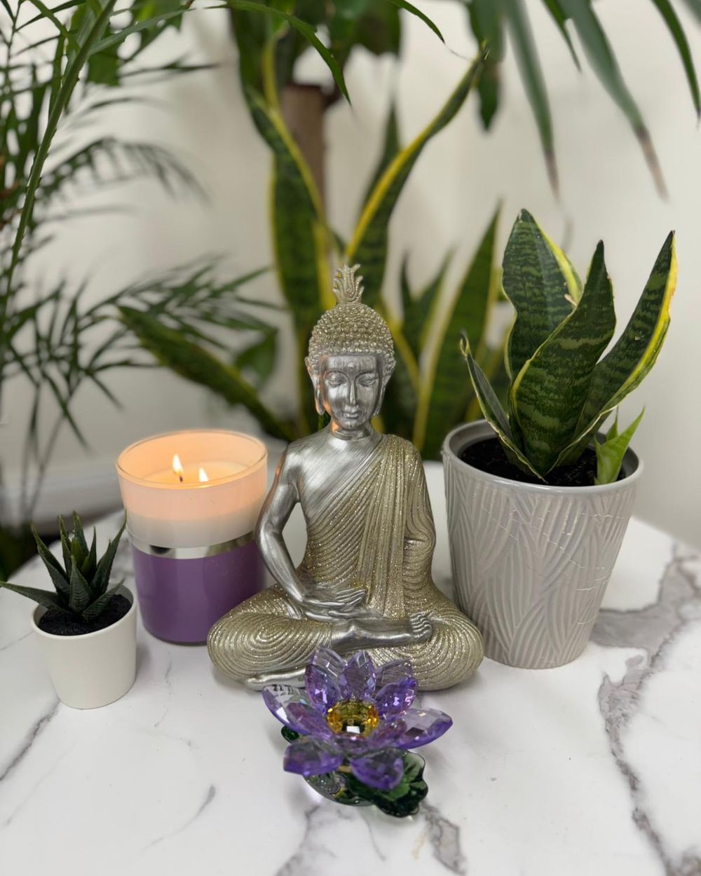 A silver Buddha on the altar with a lit candle, a glass lotus, and plants