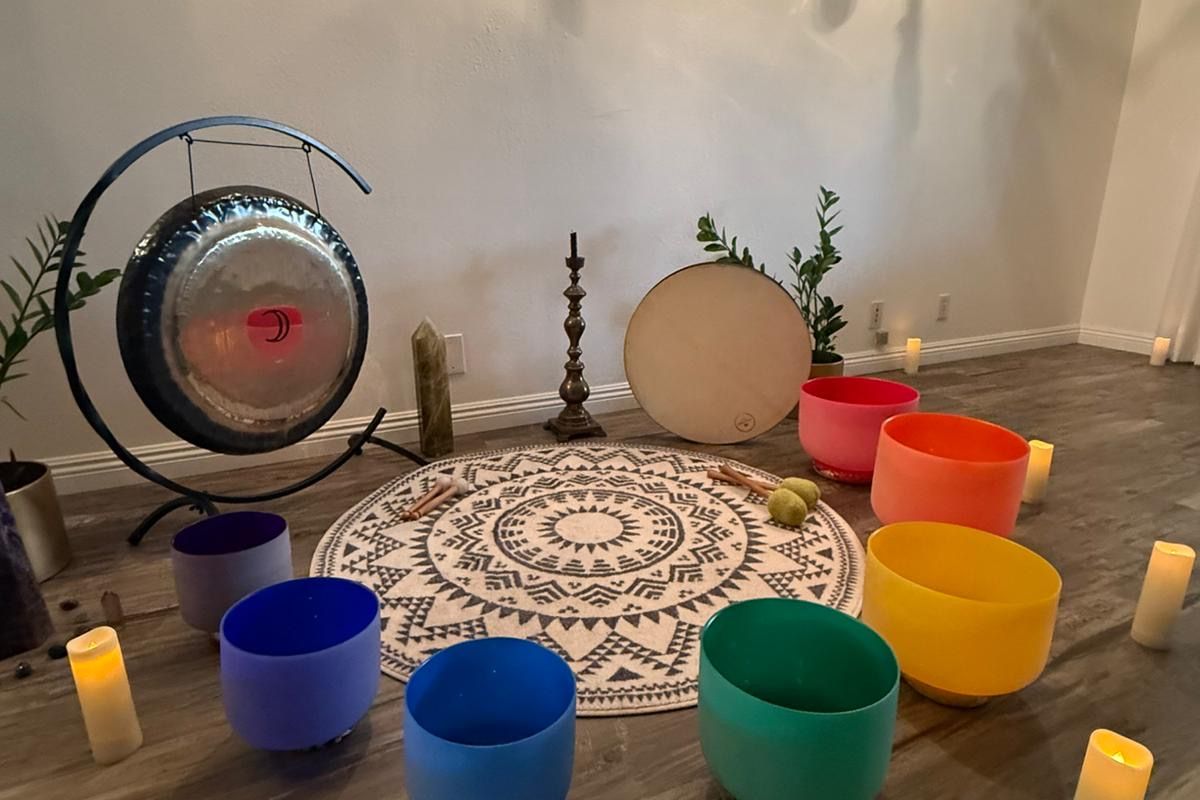 The Healing Sanctuary set for a sound bath — a full gong, a frame drum, and a circle of chakra crystal singing bowls on a mandala rug, lit by candles